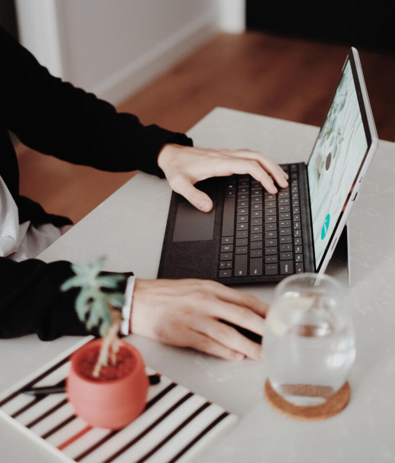 A writer typing on their laptop on a white desk, with a small plant beside them